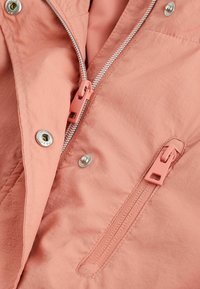 Lätt rosa jacka med en mjuk textur, silverfärgad dragkedja och knappknäppning, samt en sidoficka med dragkedja och kontrasterande metalldetaljer.