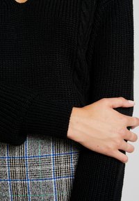 Schwarzer Strickpullover mit V-Ausschnitt, der über ein Zopfmuster an den Ärmeln und gerippte Bündchen verfügt. Kombiniert mit einem Rock im karierten Muster.