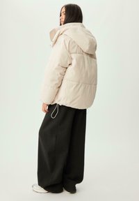 Puffjacka i ljusbeige med huva, med en fyrkantig form och elastiska snören i fållen. Matchad med svarta vida byxor.
