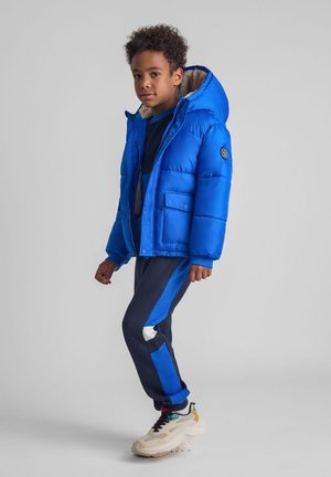 Blauwe pufferjack met een capuchon, voorzien van een nepbont voering en voorzakken. Gecombineerd met marineblauwe sweatpants en chunky witte sneakers.
