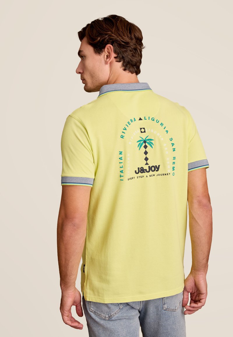 Polo shirt jaune avec un col et des poignets contrastants en bleu. Le dos présente un design graphique avec un palmier et du texte sur San Remo.