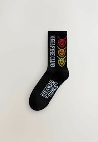 Sorte crew-sokker med hvidt "STRANGER THINGS"-tryk og farverige dragehoveder i rød, orange og gul langs siden.