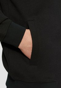 Schwarzer Sweatshirt mit Struktur, ausgestattet mit einem gerippten Bund und einer seitlichen Tasche mit horizontalem Eingang. Nahaufnahme einer Hand in der Tasche.