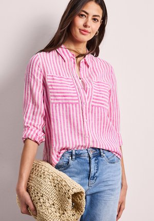 Vrouw draagt een roze-wit gestreept overhemd met knopen en lichtblauwe spijkerbroek, houdt een grote gevlochten stro clutch tas naast zich vast.