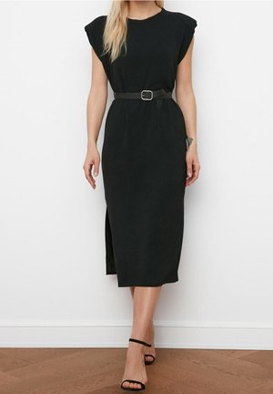 Kvinde iført en sort ærmeløs midikjole med polstrede skulderpuder, et sort bælte og sorte ankelsandaler med strop, stående op ad en hvid væg.