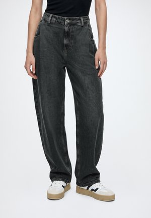 Femme portant un jean noir ample taille haute et des baskets blanches avec des détails noirs et beiges, debout devant un fond uni.