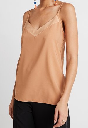 Femme portant un caraco en satin beige sans manches avec un col en V et des fines bretelles, associé à un pantalon noir et des boucles d’oreilles colorées.