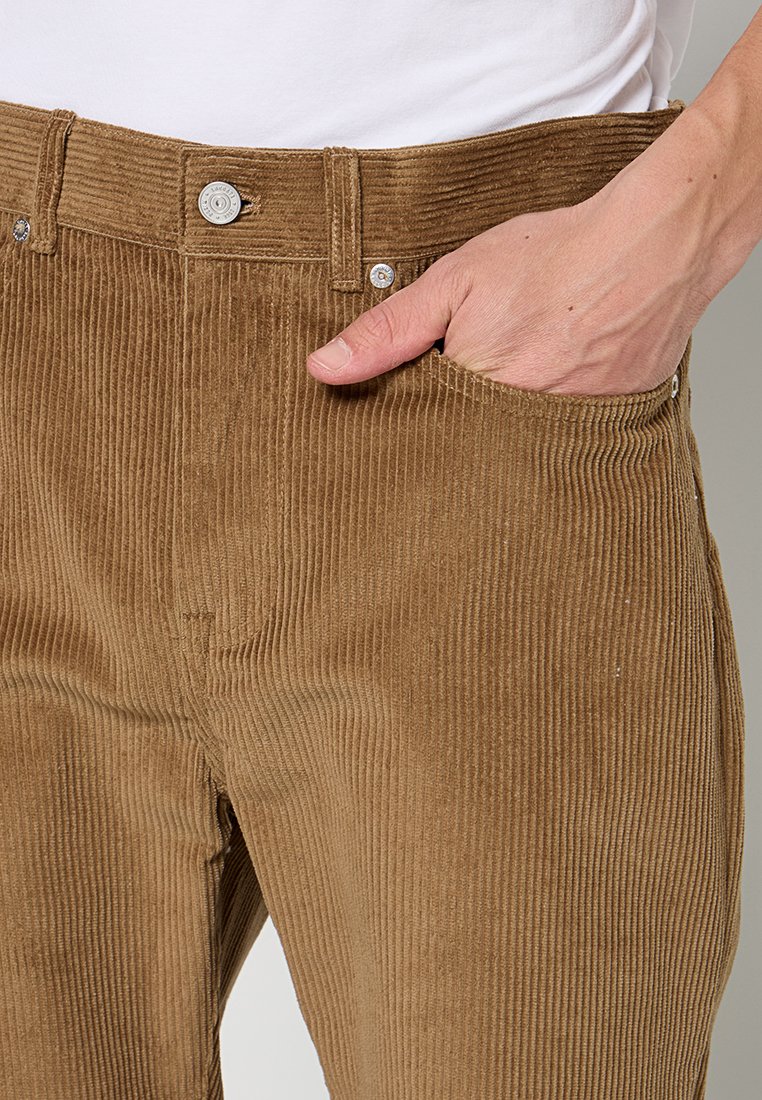Pantalons en velours côtelé marron avec une texture à côtes verticales, présentant une poche avant et des accents de boutons en métal argenté à la taille.