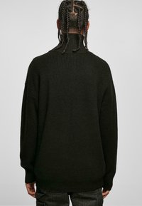 Pull en maille noir avec un col montant, texture côtelée, coupe oversize, épaules tombantes et manches longues. Détails de couture subtils visibles.