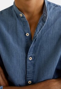 Chemise bleue sans col avec de fines rayures blanches verticales, boutonnée avec des boutons marron clair, portée par une personne aux bras croisés.