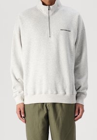 Sudadera gris con cremallera y cuello alto, puños y dobladillo acanalados. Presenta un pequeño logo negro en el pecho. Textura suave.