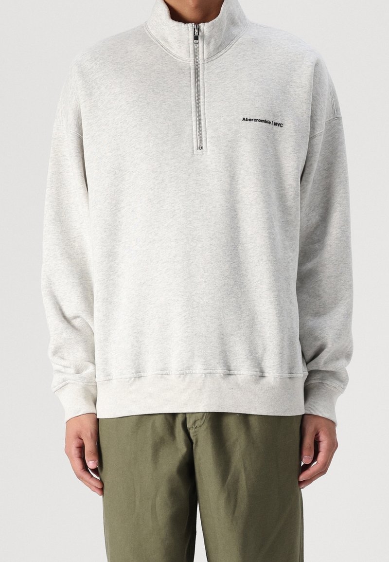 Sudadera gris con cremallera y cuello alto, puños y dobladillo acanalados. Presenta un pequeño logo negro en el pecho. Textura suave.
