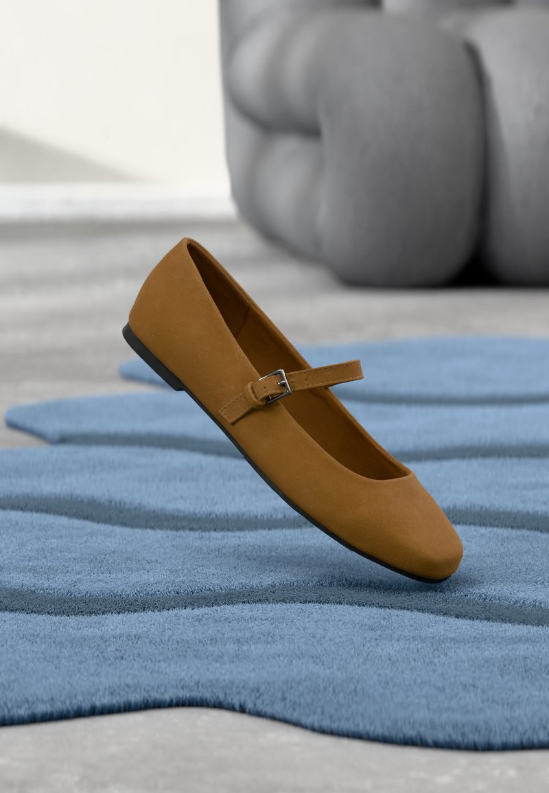 Chaussure plate Mary Jane marron avec bride à boucle flottant au-dessus d'un tapis bleu texturé, mobilier tapissé gris en arrière-plan.