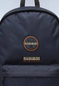 Mėlynas audinio kuprinė turi apvalų logotipą su oranžinėmis detalėmis ir stačiakampį pleistrą su prekės ženklu. Matomas užtrauktukas ir tekstūruotas paviršius.