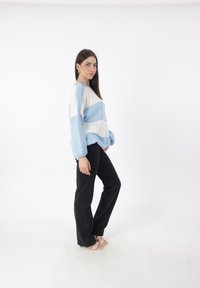 Hellblau und weiß gestreifter gestrickter Pullover mit Ballonärmeln, kombiniert mit schwarzen Schlagjeans und Riemchensandalen. Schlichter weißer Hintergrund.