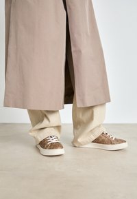 Bež trench coat kerge beeži laiade pükste peal. Pruunid tossud tekstuuriga mustriga ja valgete paeltega. Sile, minimalistlik põrand.
