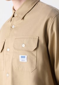 Chemise beige à boutons avec poches poitrine à rabat, boutons blancs et petite étiquette "HUGO" bleue et blanche sur la poche gauche.