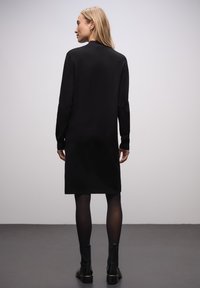 Robe noire mi-genou avec manches longues, col rond et texture douce. Portée avec des bottines noires et des collants transparents. Design simple et minimaliste.