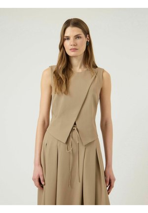 Kobieta o jasnobrązowych włosach, ubrana w bezrękawnik beżowego topu kopertowego i dopasowaną plisowaną spódnicę na jednolitym jasnym tle.