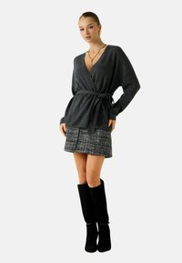 Maglione avvolgente grigio scuro con cintura annodata, abbinato a una gonna in tweed nero e bianco con trama strutturata e stivali neri al ginocchio.