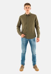 Olijfgroene overhemd met lange mouwen en knoopsluiting, gemaakt van gestructureerde stof. Gepaard met lichtblauwe jeans en beige sneakers.