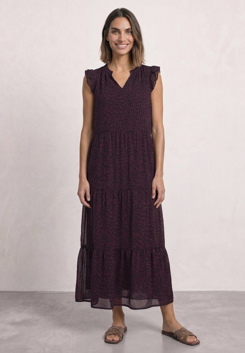 Femme souriante portant une robe midi sans manches violet foncé à motifs et des sandales plates marron, debout sur un sol gris clair avec un fond uni.