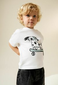 Bela bombažna majica z risbo črnega psa na deski. Otrok nosi temno sive kavbojke. Enostaven okrogel izrez in kratki rokavi.