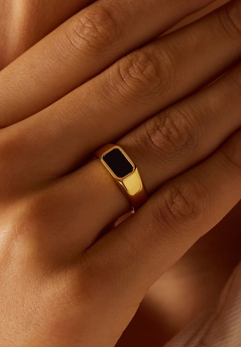 Bague en or avec une pierre noire rectangulaire portée au majeur d'une main à carnation moyenne.