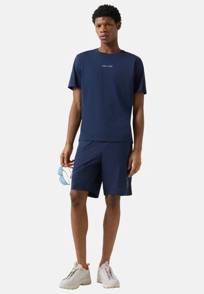 Jeune homme portant un t-shirt et un short Fire+Ice bleu marine, tenant des lunettes de soleil bleues, debout en baskets blanches sur un fond uni.