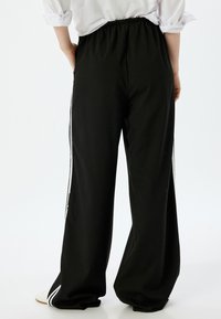 Pantalones de pierna ancha negros con cinturilla elástica. Presentan franjas laterales blancas y una textura suave. Llevado con una camisa blanca parcialmente enrollada.