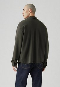 Chaqueta verde oscuro abotonada, de manga larga, ajuste holgado y costuras visibles. Combinada con vaqueros azules, mostrando un look casual.