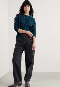 Teal pulover z vzorcem in dolgimi rokavi, v kombinaciji z visokimi črnimi džinsami in rjavim pletenim pasom. Model stoji v nevtralnem okolju.