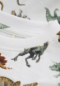 Hvidt stof t-shirt med tryk af farverige dinosaurer, ribbet krave og blød tekstur. Illustrationerne inkluderer forskellige dinosaurarter.