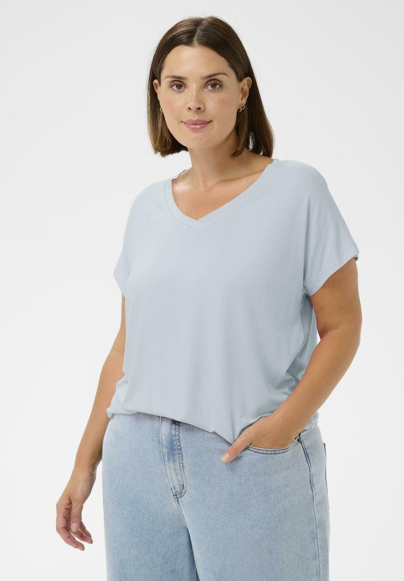 Femme aux cheveux bruns lisses portant un T-shirt bleu clair à col en V et un jean bleu clair, posant avec une main dans sa poche.