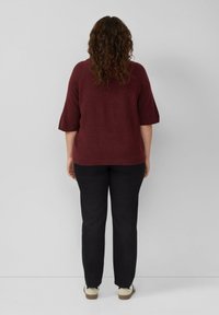 Bordeauxfarbener Strickpullover mit kurzen Ärmeln, kombiniert mit schwarzen Hosen und hellen Schuhen. Die Textur wirkt weich und lässig.