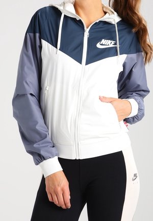 Mujer con chaqueta Nike con cremallera en blanco, azul marino y gris con capucha y leggings Nike en blanco y negro, de pie con una mano en el bolsillo.
