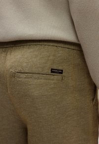 Πράσινο, υφασμάτινο παντελόνι με πίσω τσέπη και μια μικρή μαύρη ετικέτα που γράφει "SCOTCH & SODA." Το υλικό φαίνεται ελαφρύ και καθημερινό.