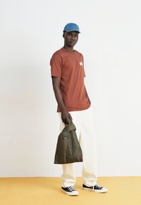 T-shirt marron avec logo, pantalon en toile large crème, sac matelassé vert et baskets noires. Casquette en denim bleu portée légèrement en angle.