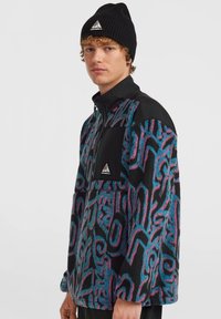Veste en polaire avec un motif abstrait noir, bleu et rose ; comporte un zip quart, des accents en panneau noir et un logo en triangle. Bonnet noir.
