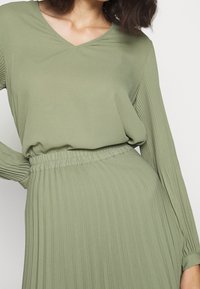 Vestito verde oliva con scollatura a V, caratterizzato da maniche lunghe e plissettate e vita arricciata; tessuto morbido con una leggera lucentezza.