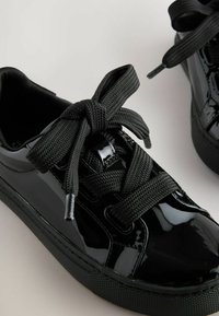 Schwarze Lackledersneaker mit mattschwarzen Stoffsenkeln und strukturiertem Gummisohle. Mit abgerundetem Zehenbereich und gepolstertem Zungenbereich.