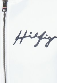 Sudadera blanca con cremallera que presenta un logo cursivo en azul oscuro que dice "Hilfiger". Cremallera plateada expuesta con un pequeño detalle de marca en el tirador de la cremallera.