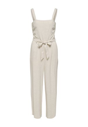 Femme aux longs cheveux foncés portant une combinaison beige sans manches avec ceinture et des mocassins beiges, debout avec une main dans la poche.
