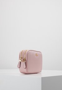 Pieni neliömäinen vaaleanpunainen nahkainen kosmetiikkalaukku, jossa on kaksi kultasta vetoketjua ja nahkasta vedettävää, joka on hienovaraisella tekstuurilla ja kohokuvioidulla logolla.