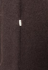 Brun texturerad tyg med en mjuk, lurvig yta; har en liten vit etikett med "MAIRZ" broderat på sidan.