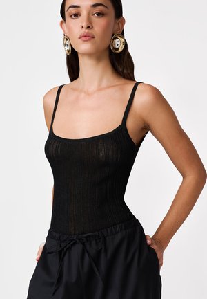 Kobieta ubrana w czarny prążkowany top na ramiączkach, czarne spodnie ze ściągaczem i duże, okrągłe złote kolczyki na jednolitym tle.