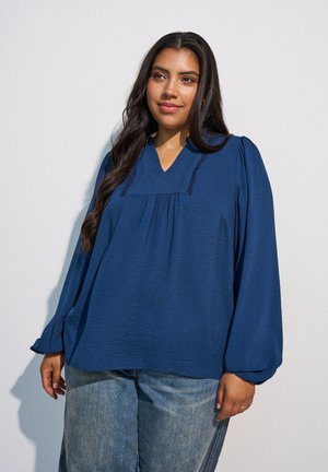 Kvinde med langt mørkt hår iført en løs mørkeblå bluse med lange pufærmer og lyseblå jeans, der står mod en ensfarvet baggrund.