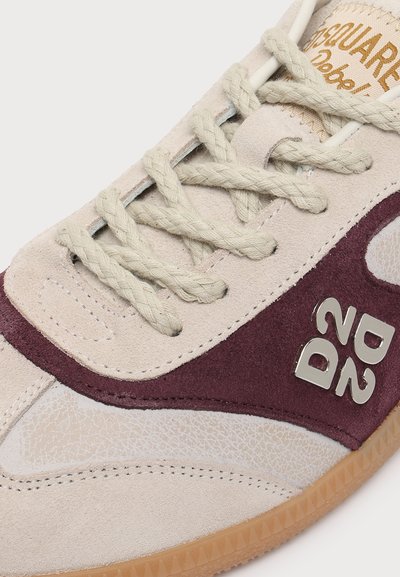 Gros plan sur une basket avec des lacets beiges, des empiècements en suède bordeaux et crème, une semelle en gomme et un logo métallique "DSQ2" sur le côté.