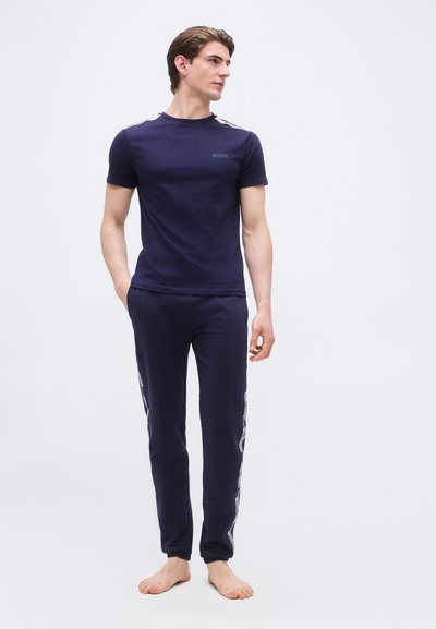 Homme debout pieds nus portant un t-shirt à manches courtes bleu marine et un pantalon assorti avec des bandes de texte blanches sur les côtés.