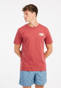 Raudona medvilninė t-shirt marškinėliai su trumpomis rankovėmis ir apvalia iškirpte, kuriuose kairėje krūtinės pusėje yra mažas baltas logotipas. Derinama su šviesiais mėlynais šortais, puoštomis palmėmis.
